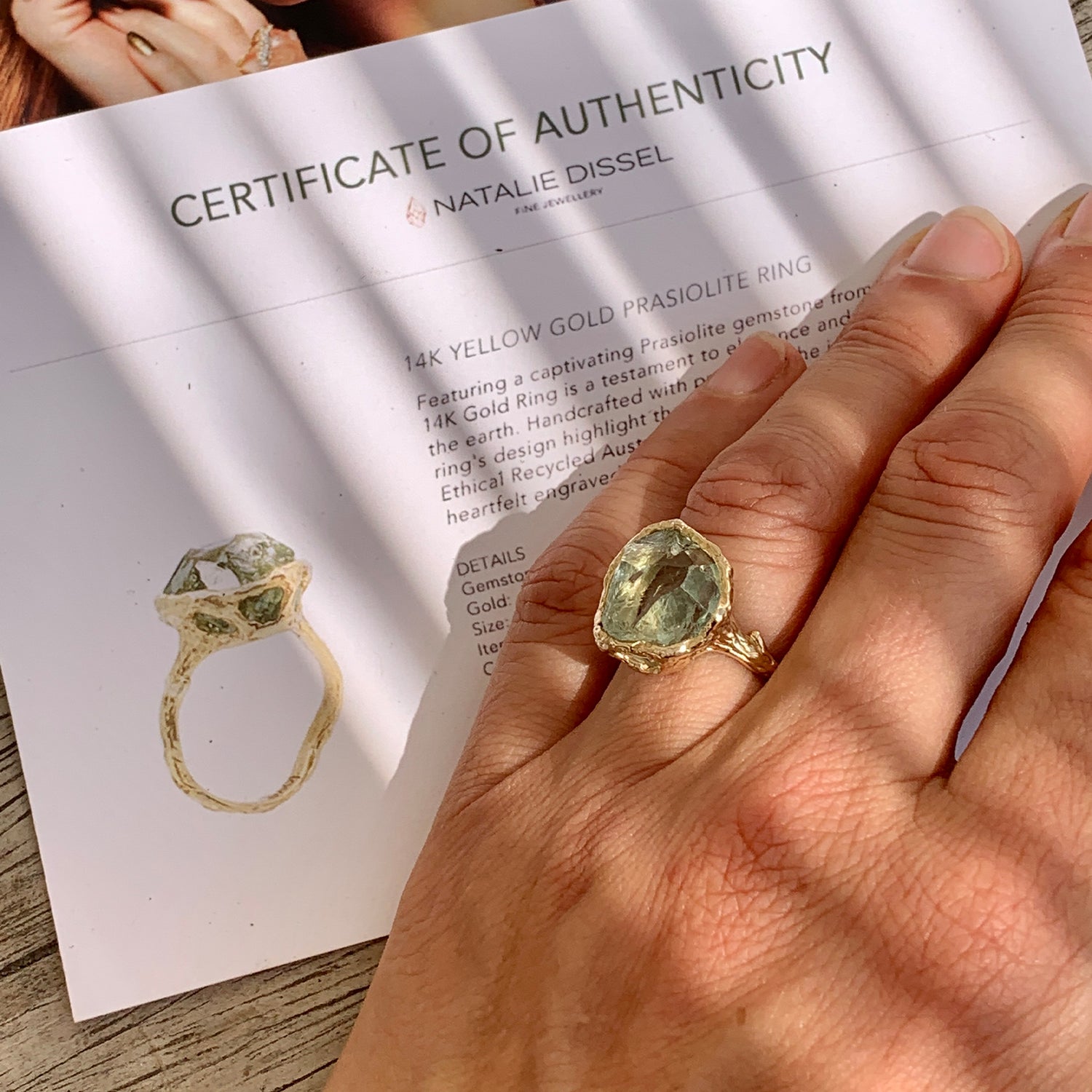 Gold ring with green gemstone prasiolite on a hand, next to a certificate of authenticity for the ring by natalie dissel
