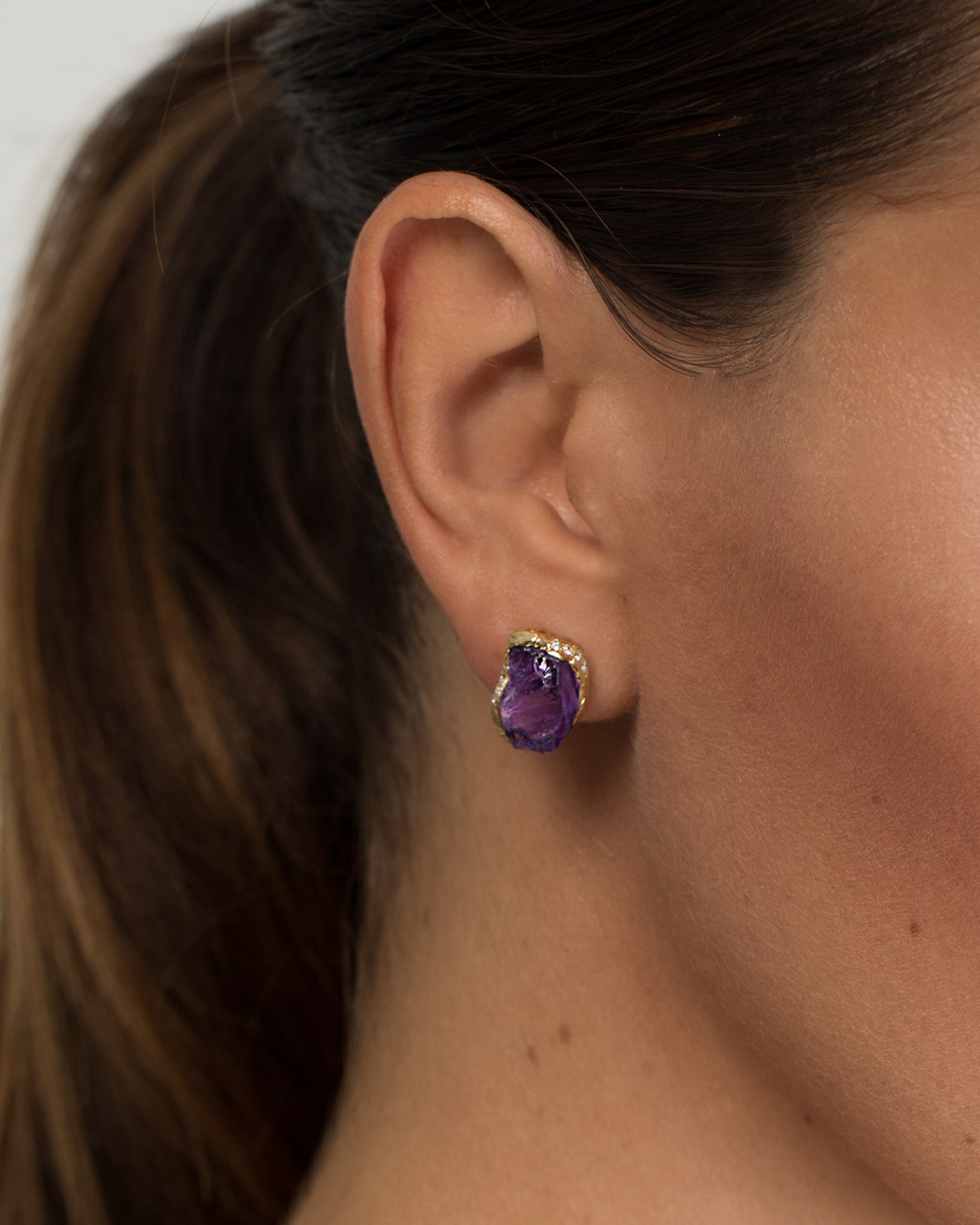 Close-up of a model wearing an organic, rough-cut purple amethyst stud earring wrapped in 18K gold and trimmed with small diamonds.