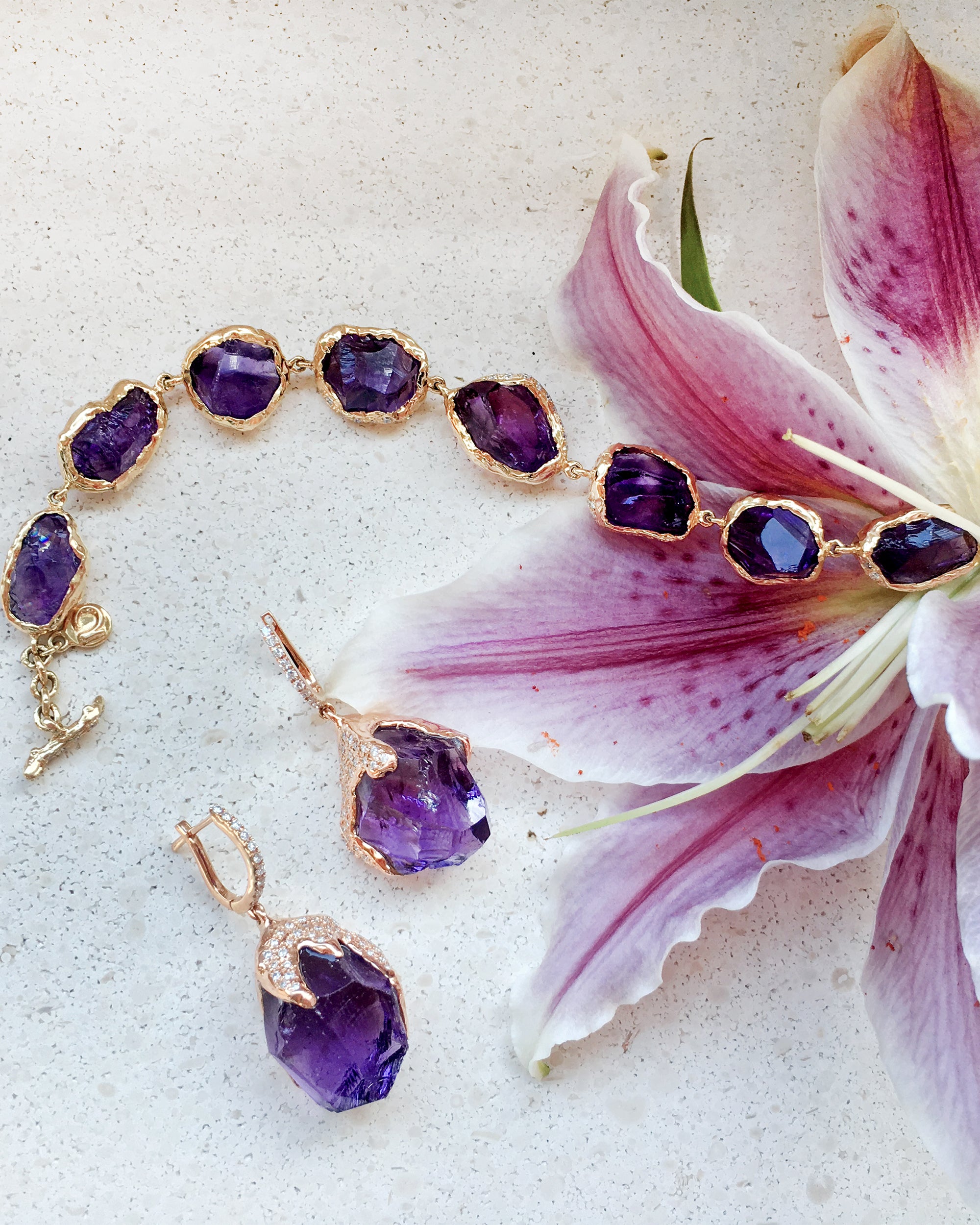 Natalie Dissel 18K yellow gold Raw Natural Amethyst gemstone link bracelet and matching Amethyst drop earrings displayed alongside a lily flower, Gem Rocks collection