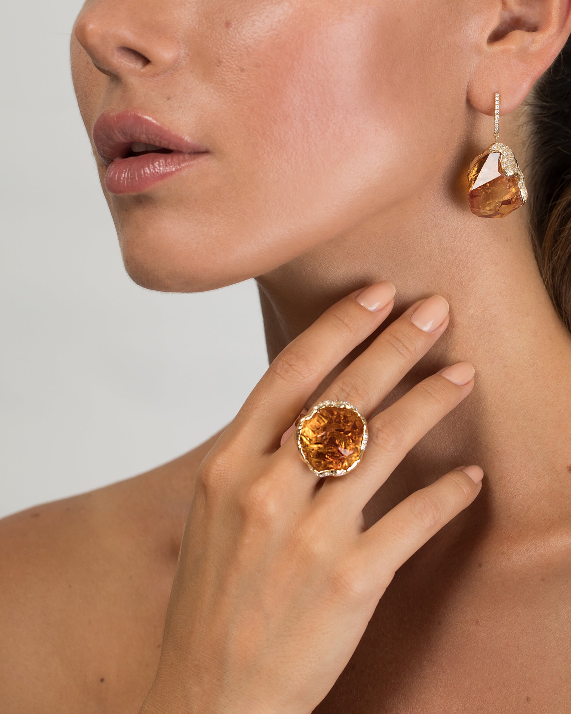 Portrait shot of a woman resting her hand on her collarbone, highlighting a bold, organic-shaped 18K gold ring with a large raw citrine gem bordered by pavé-set diamonds