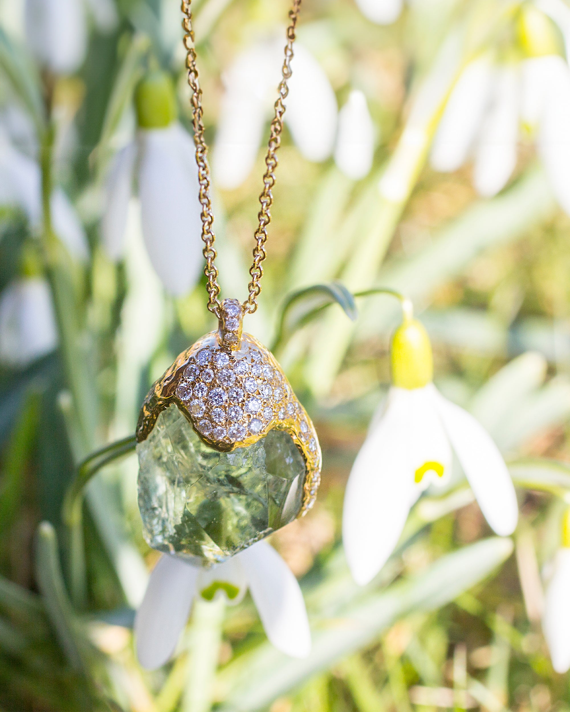 Close-up of Natalie Dissel 18K yellow gold pendant featuring a raw Prasiolite gemstone with a pavé diamond cap, set against white snowdrop flowers.