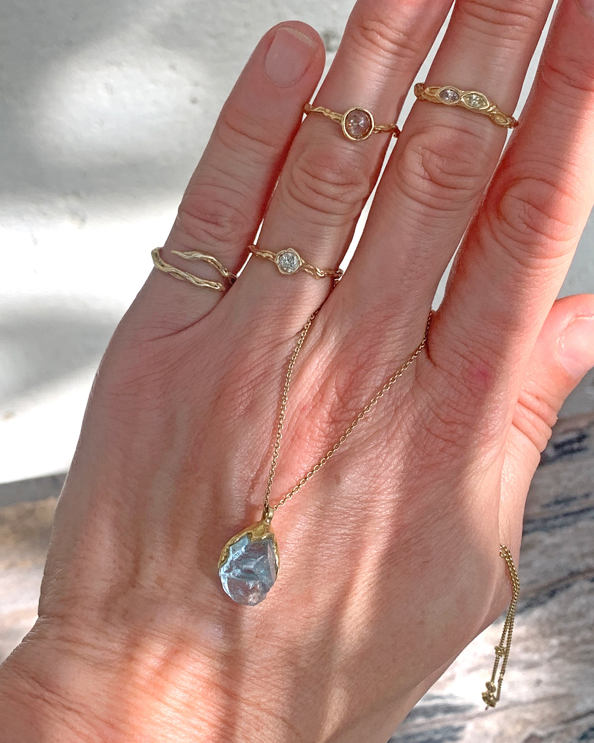 Natalie Dissel 18K yellow gold raw Sky Blue Topaz pendant necklace draped over a hand wearing delicate gold gemstone rings.